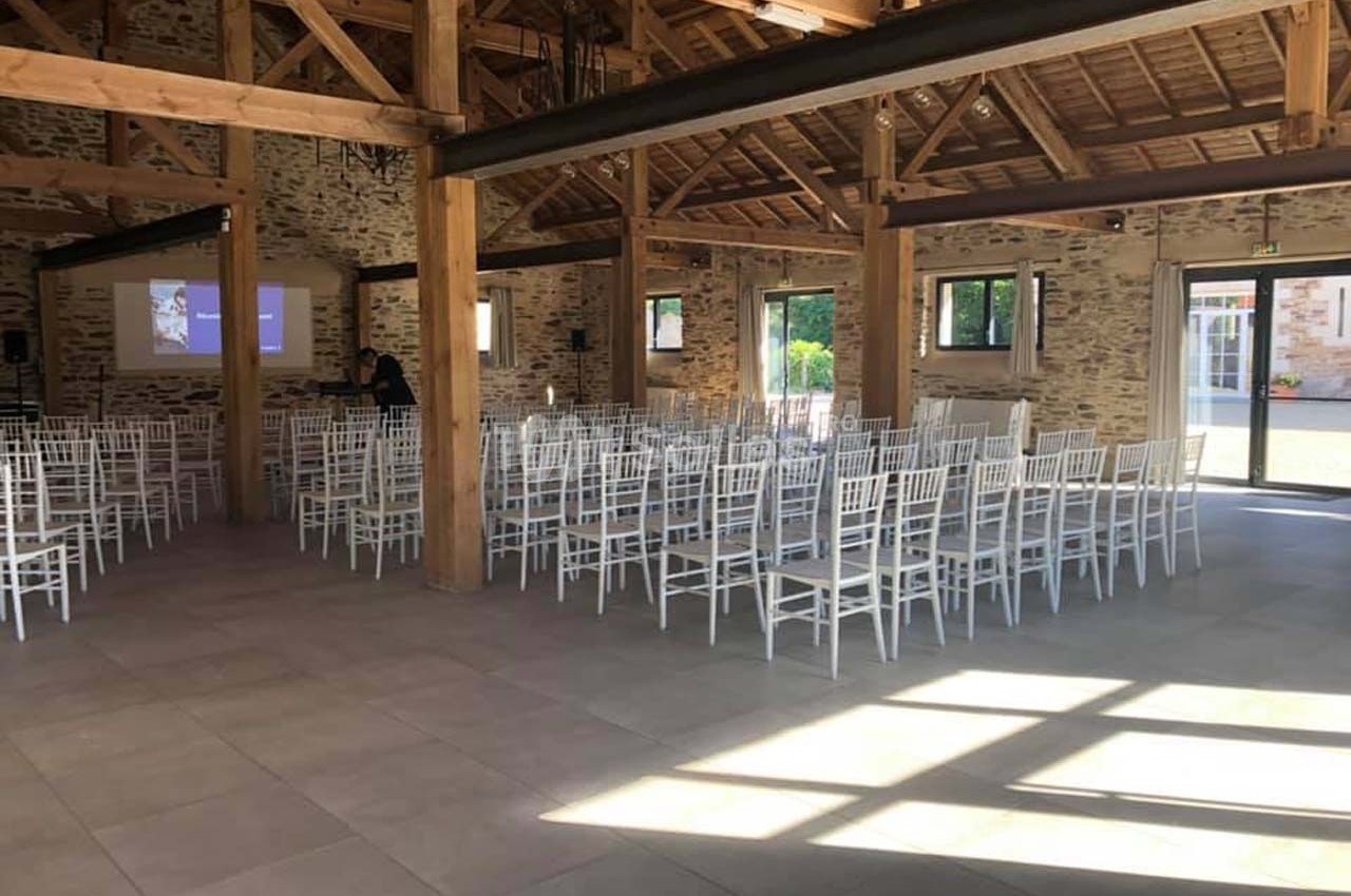 Salle de réception lumineuse avec des chaises blanches alignées et un écran de projection en arrière-plan.