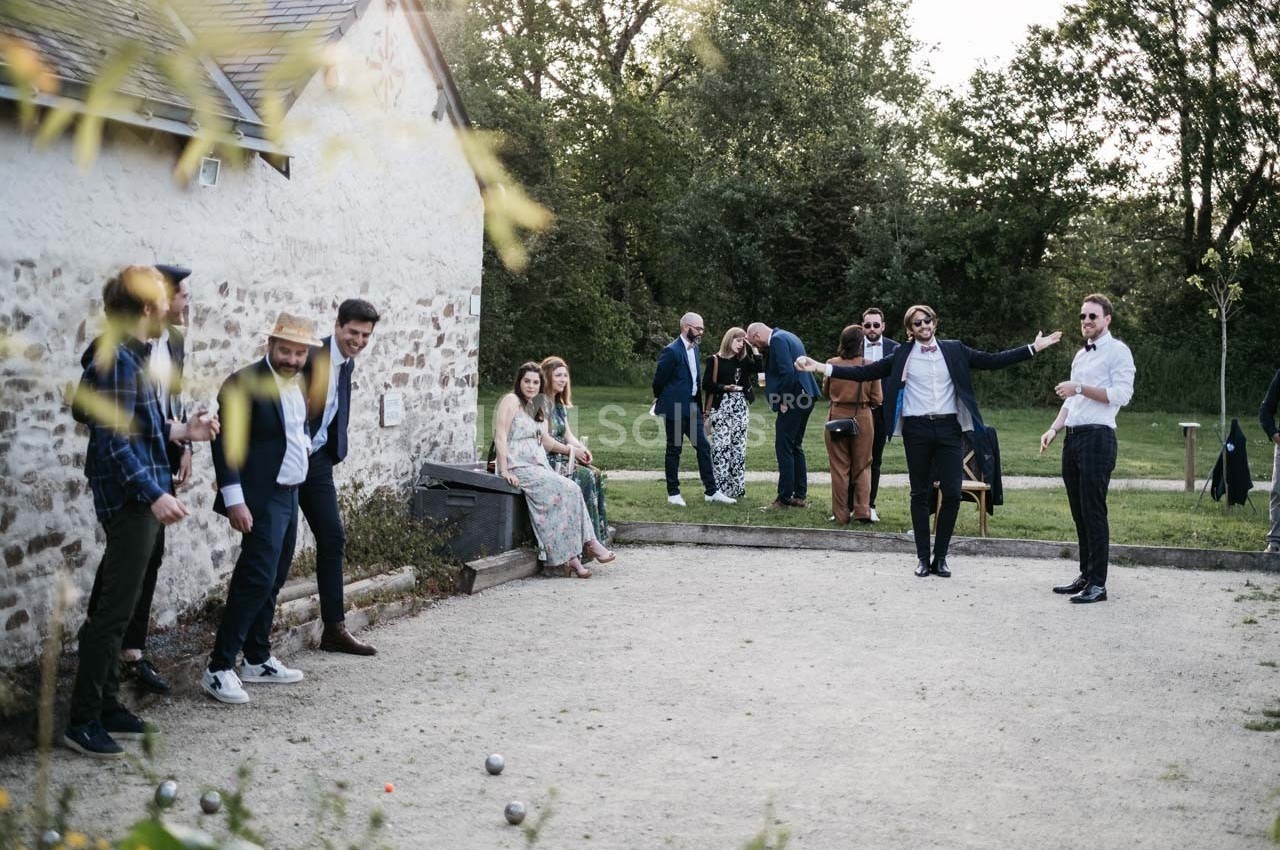 Des personnes jouent à la pétanque dans un jardin, tandis que d'autres discutent près d'une maison en pierre.