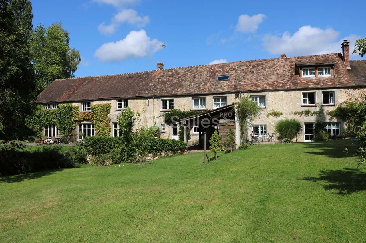 Grande maison en pierre avec toit en tuiles, entourée de verdure et d'un jardin sous un ciel bleu.