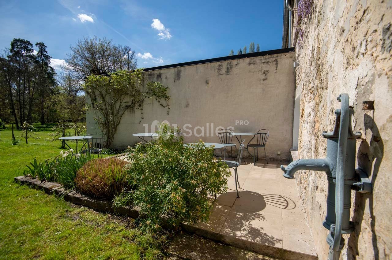 Terrasse extérieure avec tables et chaises en métal, entourée de verdure et adossée à un mur en pierre.
