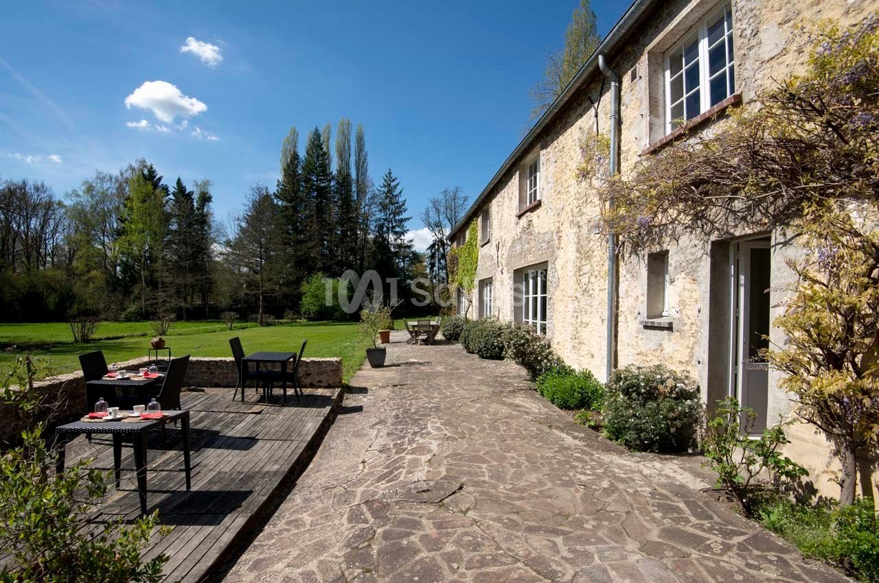 Terrasse en pierre bordée de verdure avec tables dressées, adossée à une maison en pierre sous un ciel dégagé.