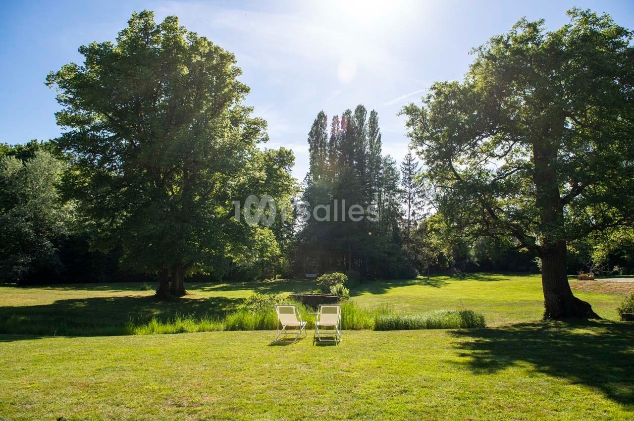 Deux chaises longues blanches sur une pelouse verdoyante entourée d'arbres sous un ciel ensoleillé.