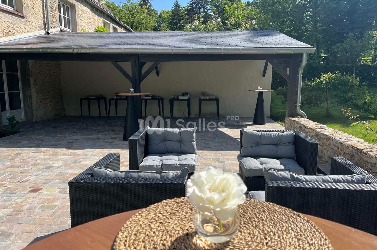 Salon de jardin en rotin avec coussins gris sur une terrasse pavée, devant un abri en bois dans un cadre verdoyant.