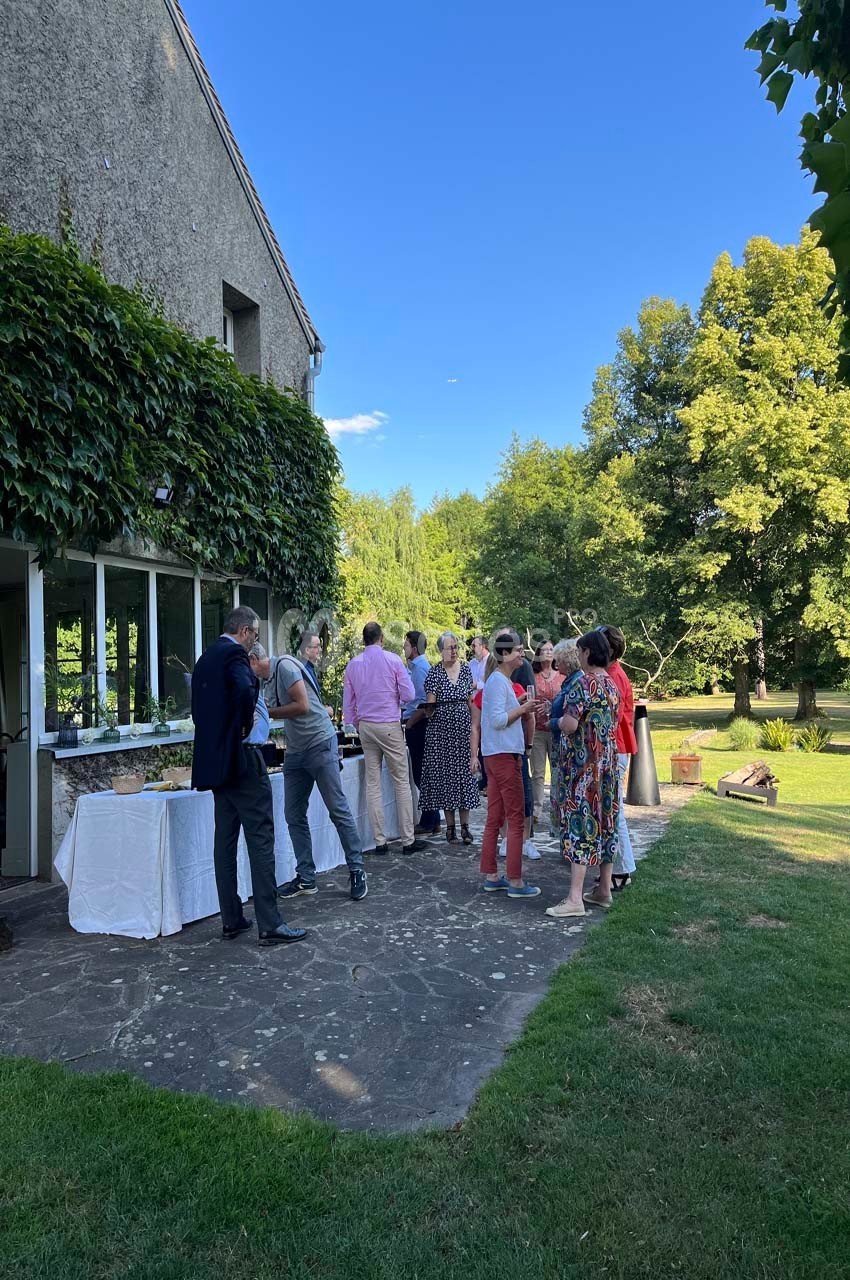 Des personnes discutent en extérieur près d'une table de buffet, devant un bâtiment couvert de végétation.