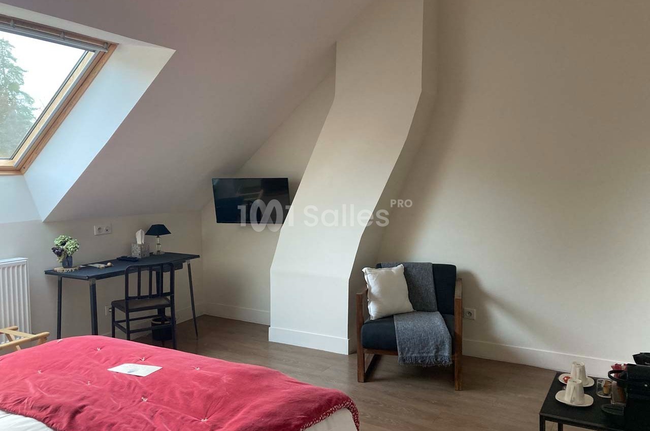 Chambre mansardée avec bureau, fauteuil, télévision murale et lit couvert d'un plaid rouge, éclairée par une fenêtre de toit.