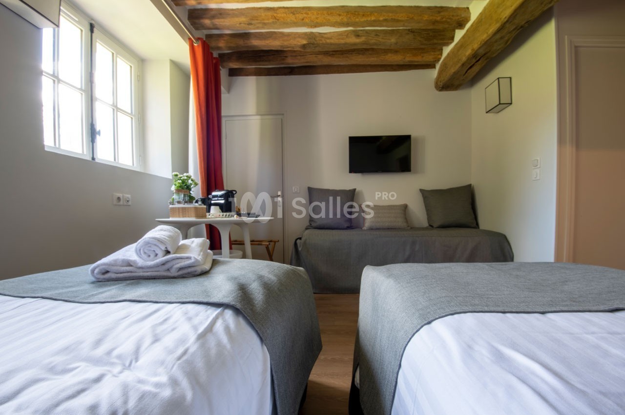 Chambre lumineuse avec deux lits simples, poutres apparentes, télévision murale et table équipée d'une machine à café.