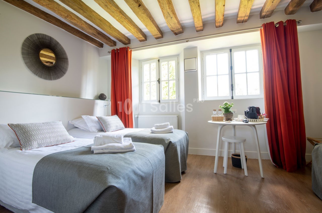 Chambre lumineuse avec deux lits simples, poutres apparentes, rideaux rouges et table avec boissons près de la fenêtre.