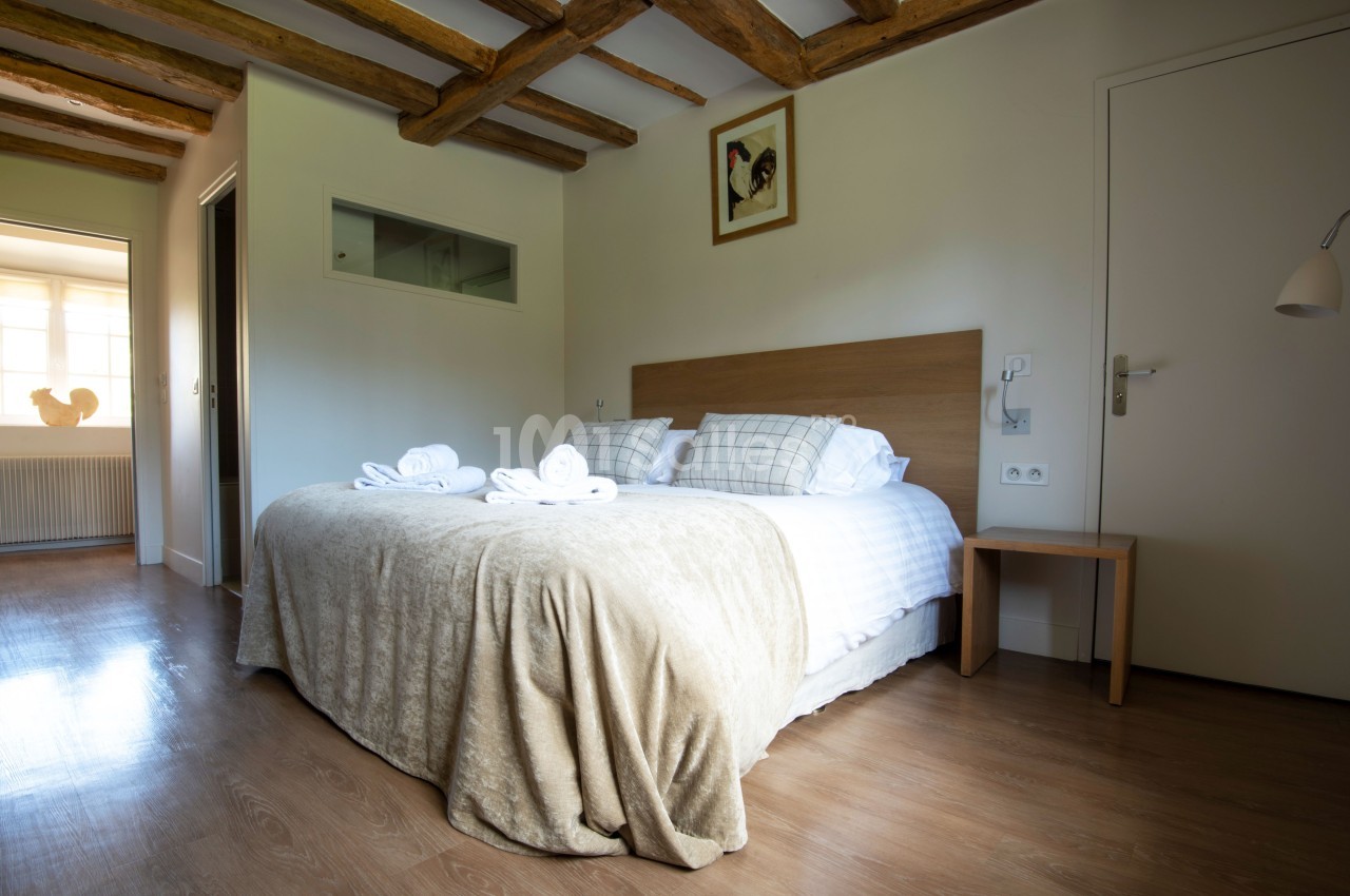 Chambre lumineuse avec lit double, draps blancs, poutres apparentes, sol en bois et décoration murale simple.