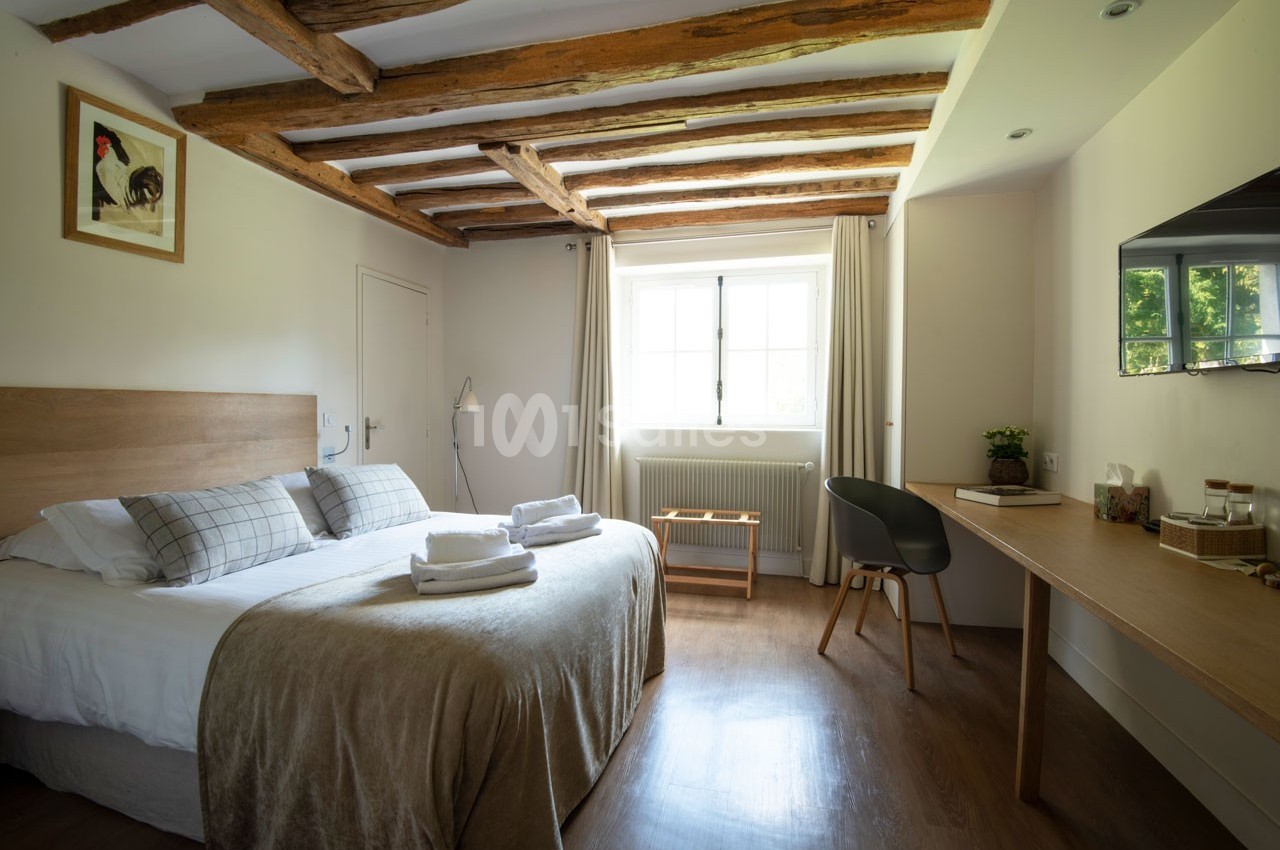 Chambre lumineuse avec lit double, poutres apparentes, bureau en bois, fenêtre donnant sur un extérieur verdoyant.