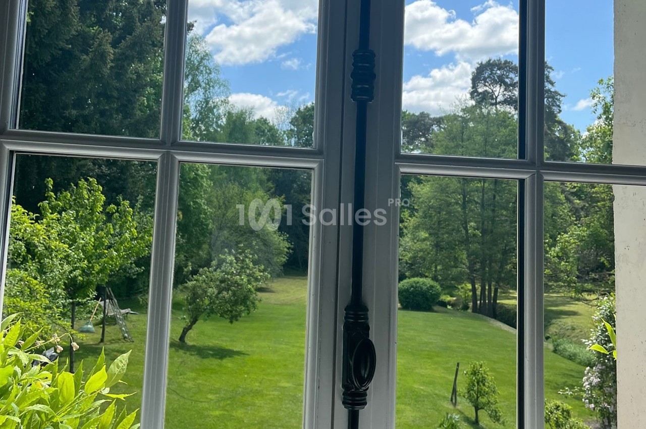 Vue sur un jardin verdoyant avec pelouse, arbres et ciel bleu, prise depuis une fenêtre à croisillons.