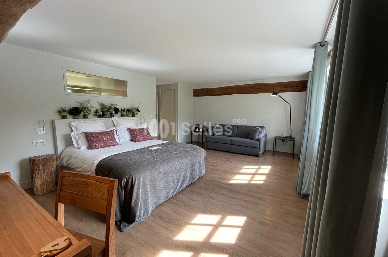 Chambre lumineuse avec lit double, canapé, bureau en bois, plantes décoratives et grande fenêtre laissant entrer la lumière.