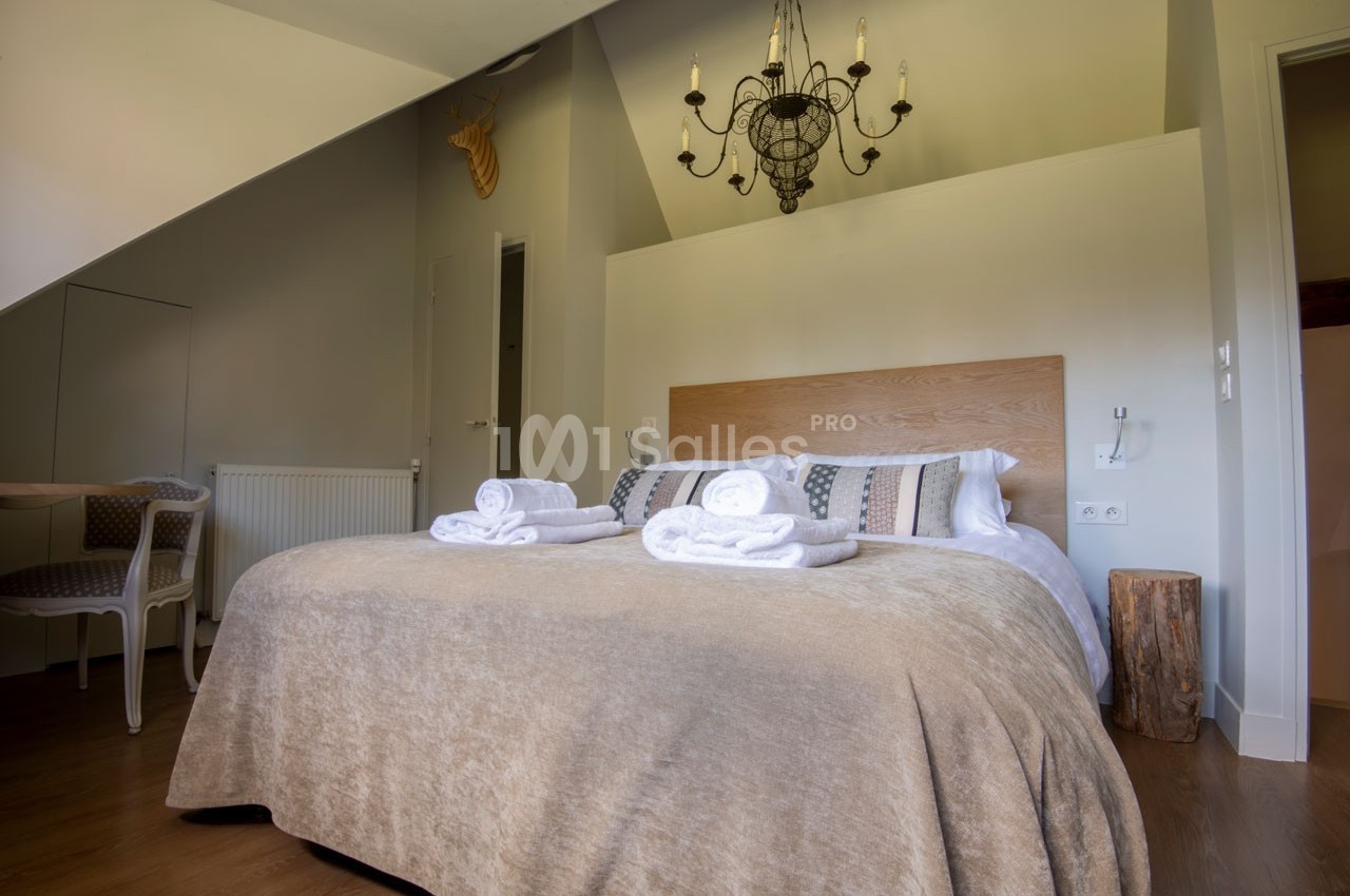 Chambre lumineuse avec lit double, linge de lit beige, serviettes pliées, lustre et décoration murale en bois.