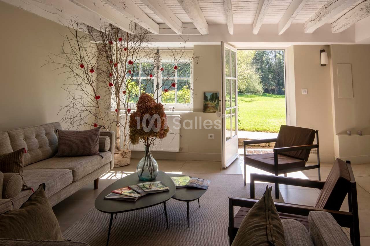 Salon lumineux avec poutres apparentes, mobilier moderne, grande baie vitrée ouverte sur un jardin verdoyant.