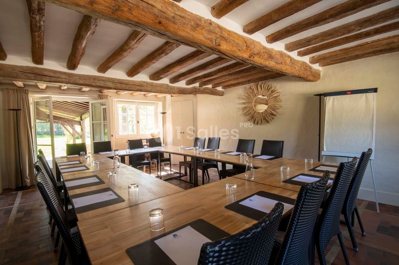 Salle de réunion lumineuse avec grandes tables en bois, chaises en osier, poutres apparentes et vue sur un jardin.