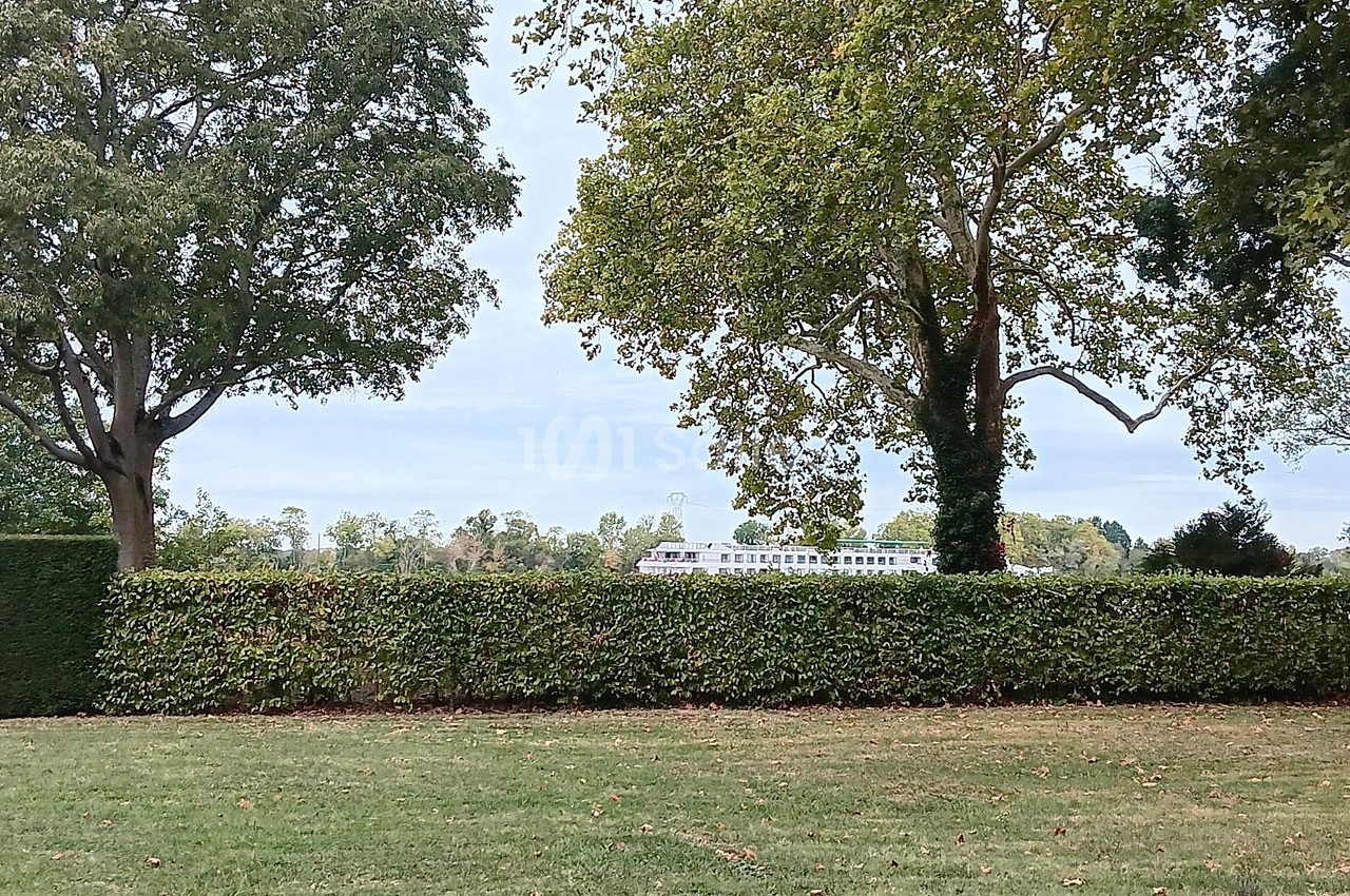 Vue d'une pelouse bordée de haies et d'arbres, avec un bâtiment blanc visible en arrière-plan.