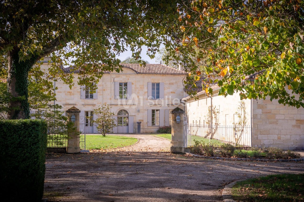 Façade d'une maison en pierre entourée d'arbres et d'un jardin, avec une allée menant à l'entrée principale.