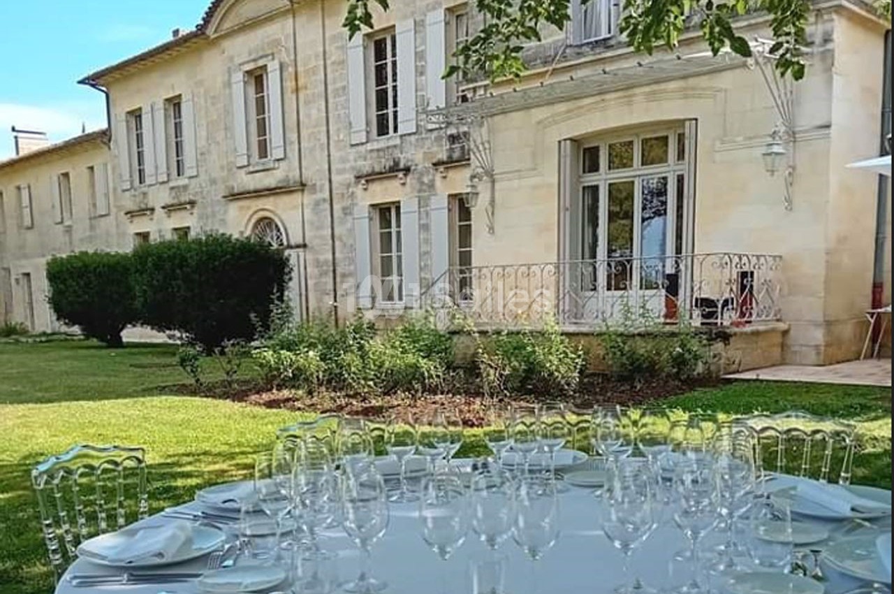 Table dressée avec verres et assiettes dans un jardin, devant une grande bâtisse en pierre avec volets blancs.