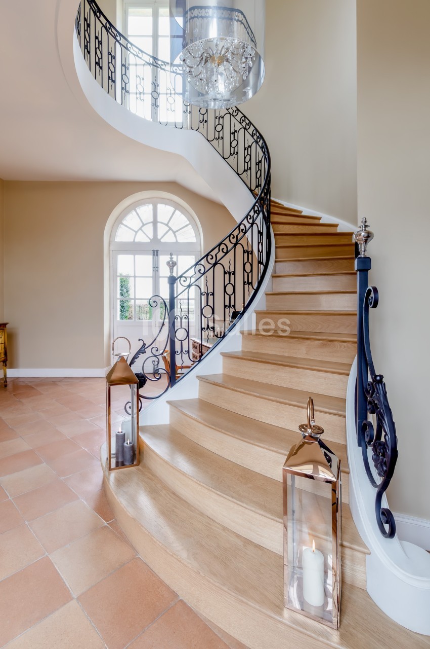 Escalier en bois courbé avec rampe en fer forgé, éclairé par une grande fenêtre et décoré de lanternes au sol.