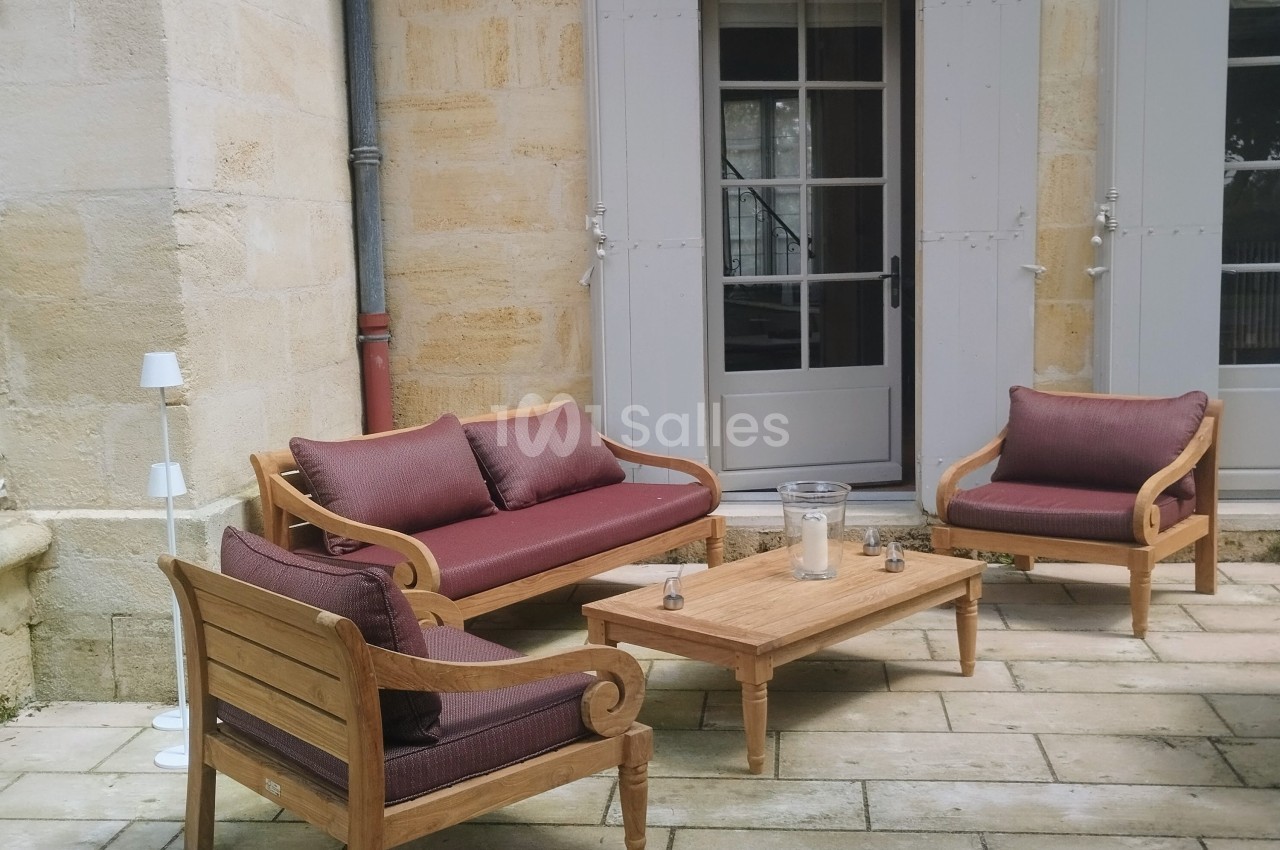 Salon de jardin en bois avec coussins bordeaux, installé sur une terrasse en pierre devant une maison aux volets gris.
