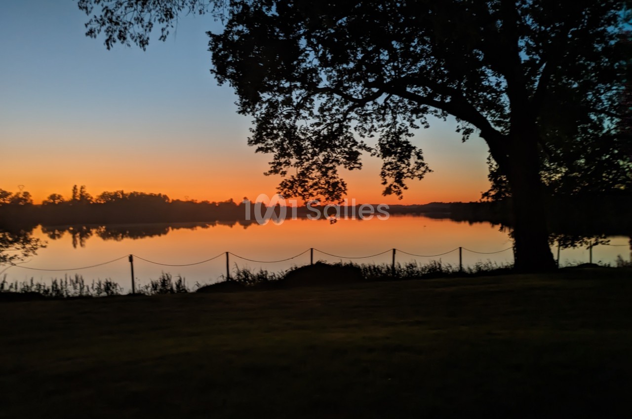 Paysage au crépuscule avec un lac calme reflétant le ciel orangé, bordé d'arbres et d'une clôture discrète.