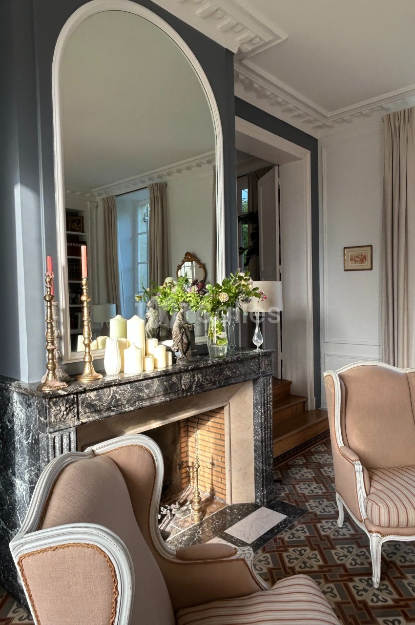 Cheminée en marbre noir décorée de bougies et de fleurs, entourée de fauteuils dans un salon lumineux.