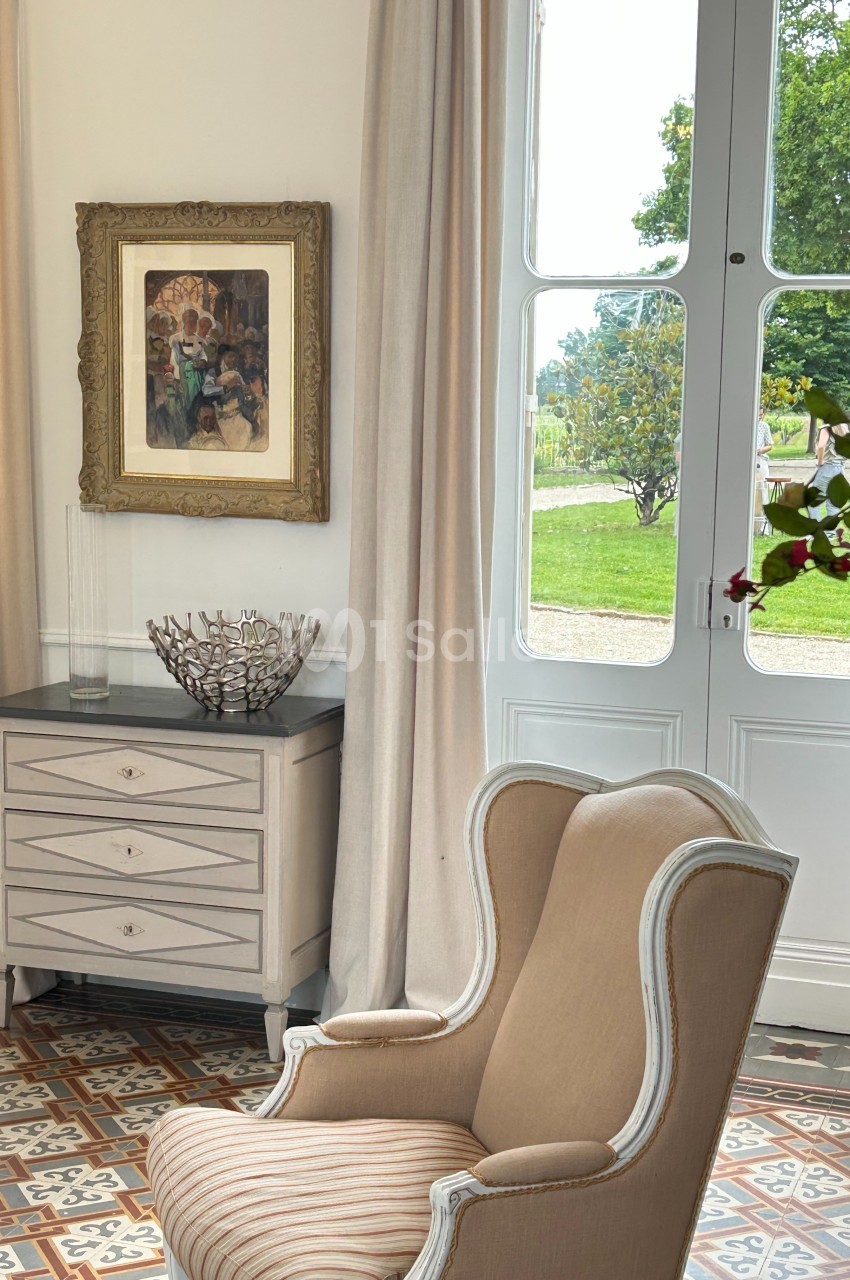 Fauteuil beige rayé devant une commode claire, près d'une fenêtre donnant sur un jardin verdoyant.