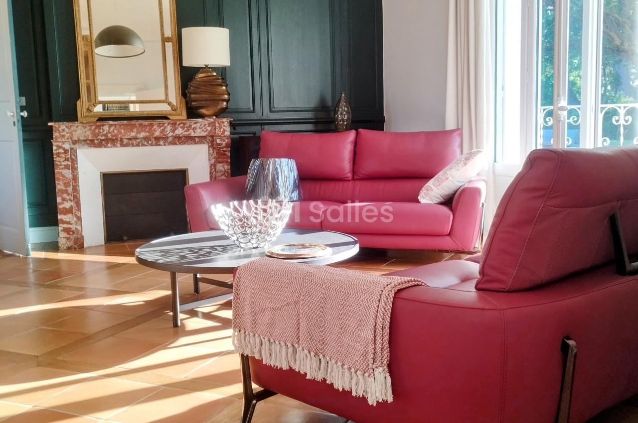 Salon lumineux avec canapé et fauteuil rouges, table basse, cheminée en marbre, miroir doré et grandes fenêtres.