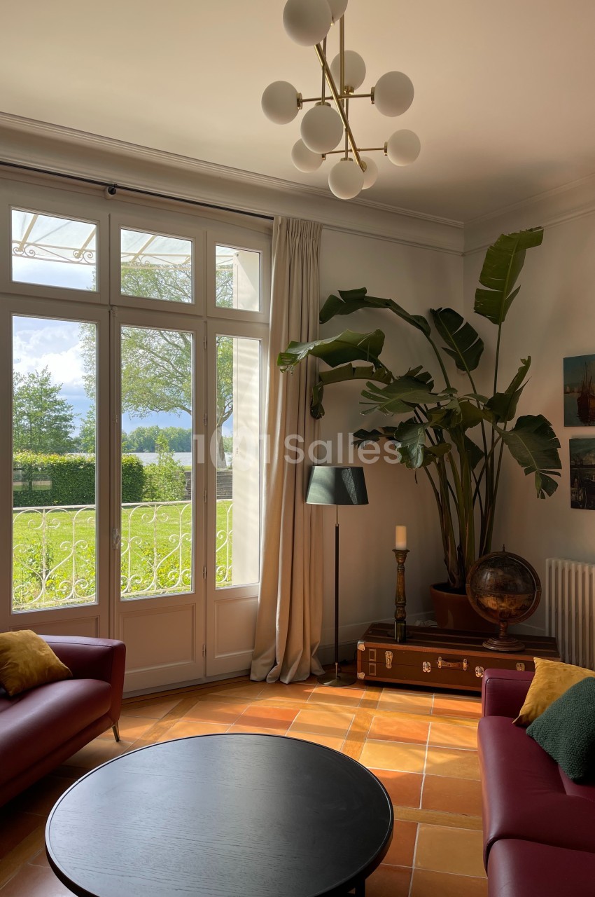 Salon lumineux avec grandes baies vitrées, mobilier moderne, plantes décoratives et vue sur un jardin verdoyant.