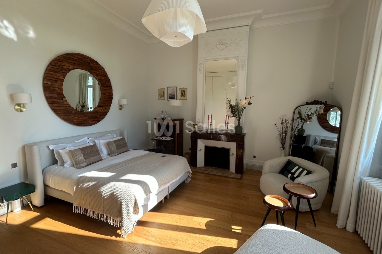 Chambre lumineuse avec lit double, miroir rond, cheminée, fauteuils et grandes fenêtres laissant entrer la lumière naturelle.