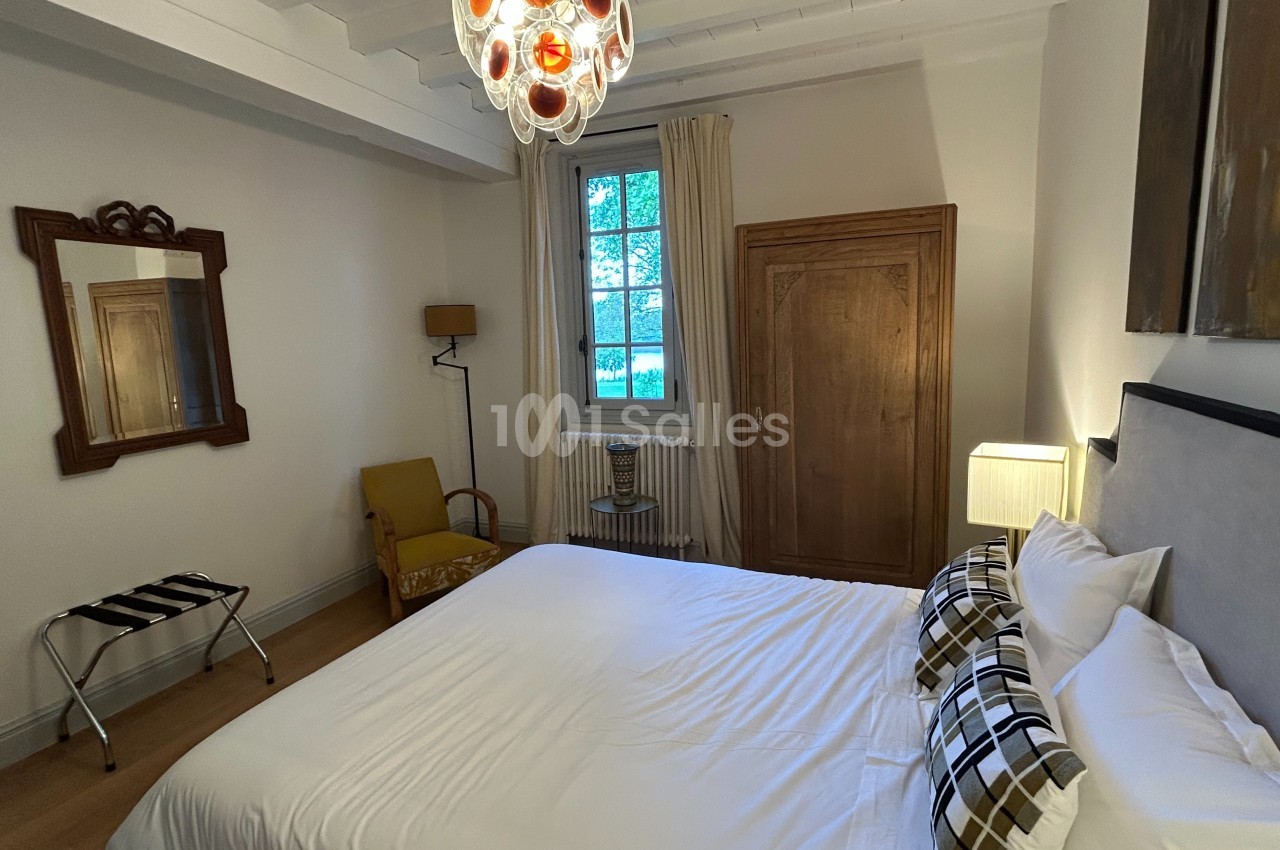 Chambre lumineuse avec lit double, armoire en bois, fauteuil jaune, miroir mural et fenêtre donnant sur un jardin.