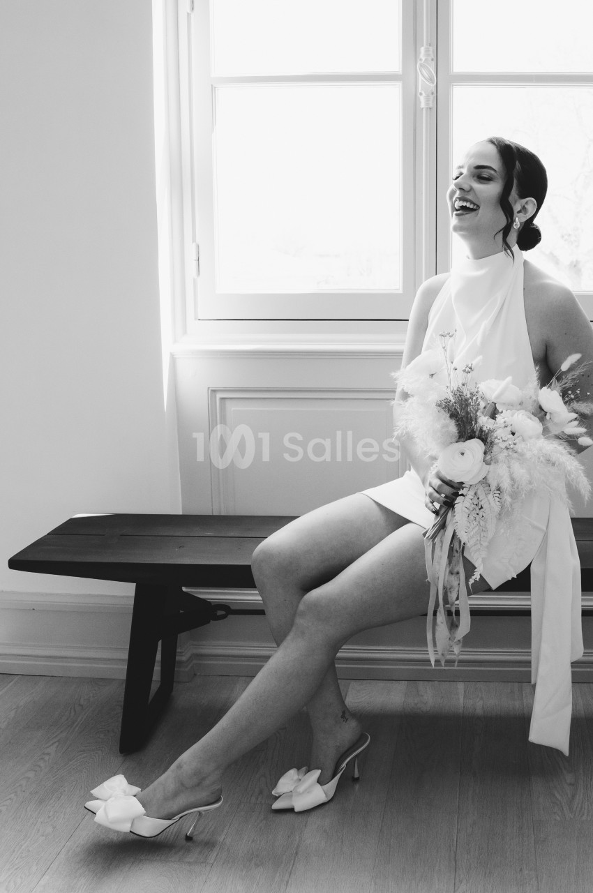 Une femme en robe blanche, assise sur un banc près d'une fenêtre, tenant un bouquet de fleurs, sourit joyeusement.