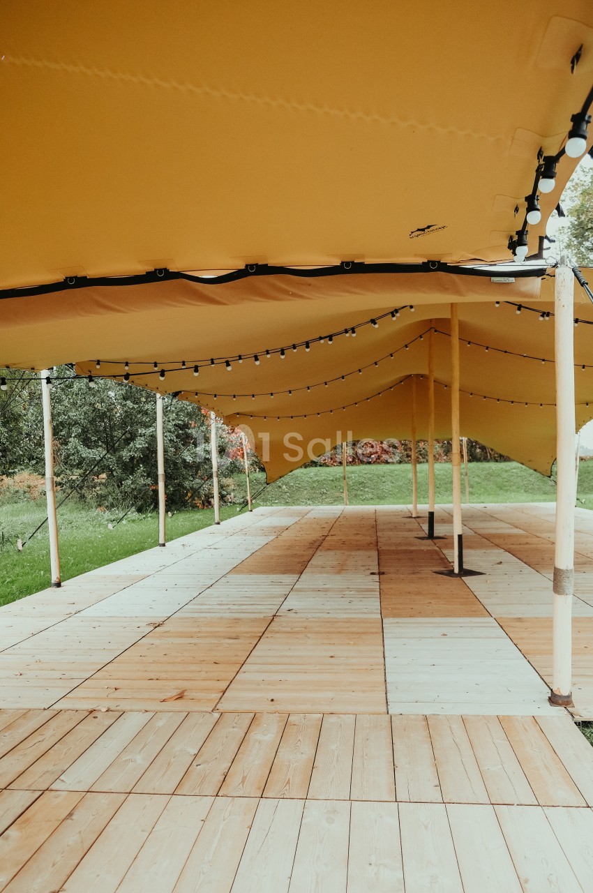 Structure en bois couverte par une toile jaune tendue, éclairée par des guirlandes lumineuses, dans un espace extérieur.