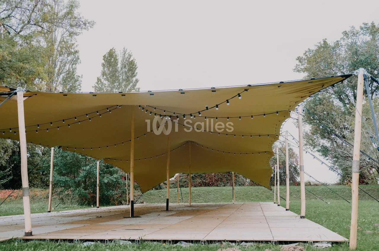 Grande tente beige tendue sur des poteaux en bois, éclairée par des guirlandes lumineuses, installée sur un sol en bois.