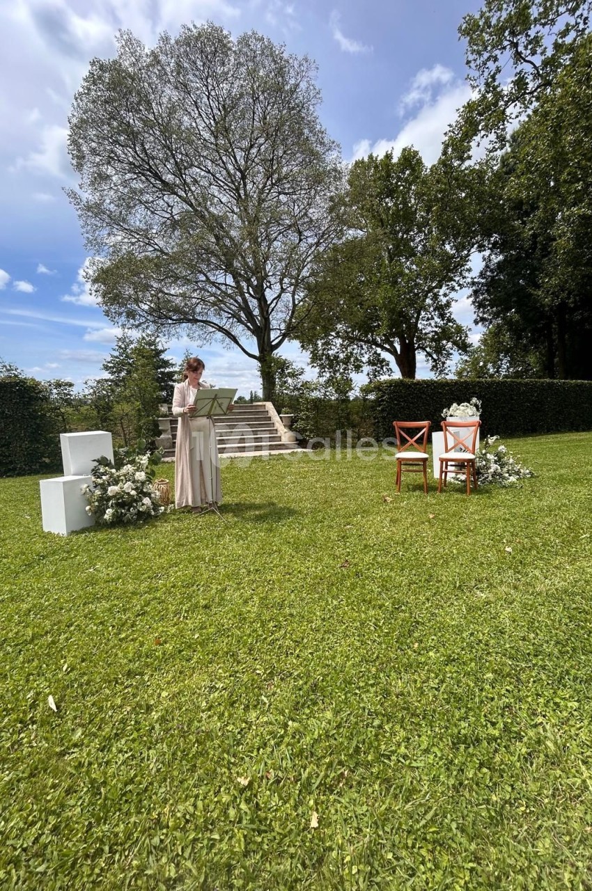 Une femme lit un texte sur un pupitre en plein air, devant des chaises décorées et un escalier dans un jardin verdoyant.