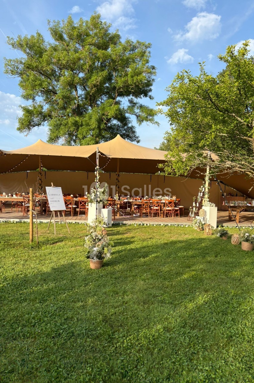 Grande tente beige installée dans un jardin verdoyant, aménagée avec des tables en bois pour un événement en plein air.