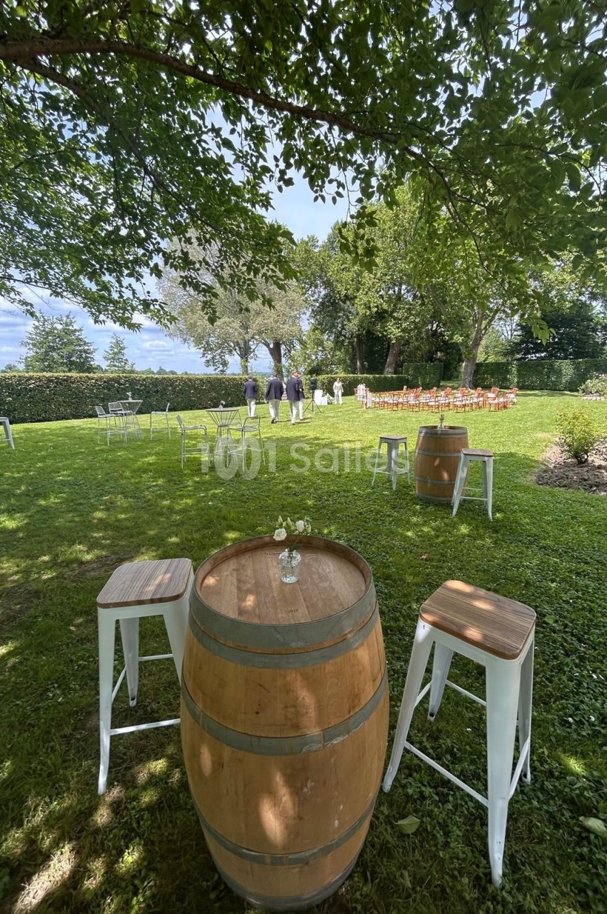 Table haute en tonneau avec tabourets dans un jardin verdoyant, disposée pour un événement en plein air.