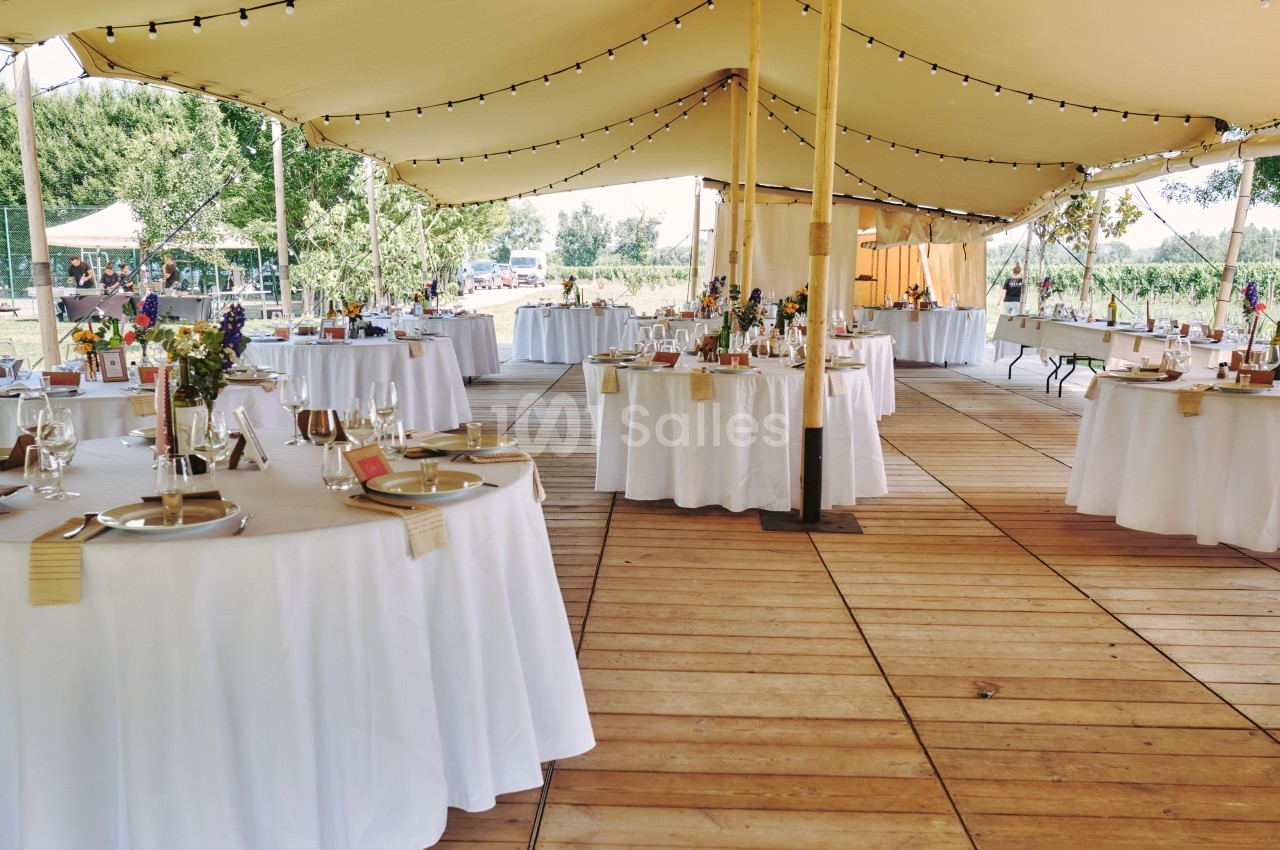 Tables rondes décorées pour un événement sous une tente avec guirlandes lumineuses, sur un sol en bois.