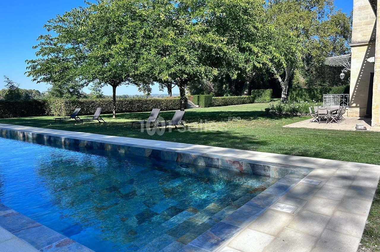 Piscine extérieure entourée de chaises longues, pelouse, arbres et terrasse avec table et chaises.