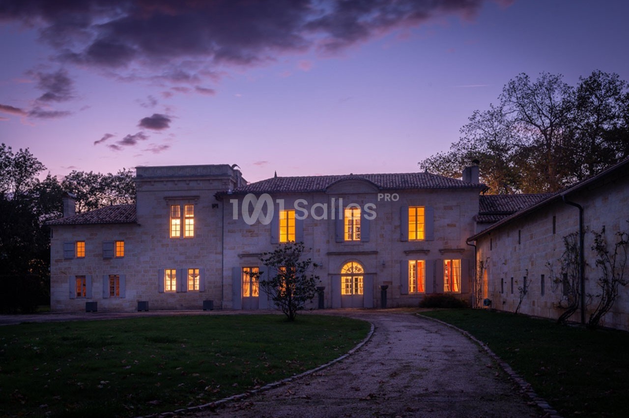 Façade d'un bâtiment en pierre éclairé de nuit, entouré d'arbres et d'une allée sinueuse.