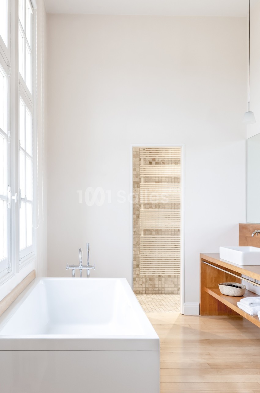 Salle de bain lumineuse avec baignoire blanche, meuble en bois, lavabo double et porte ouverte sur un sauna.