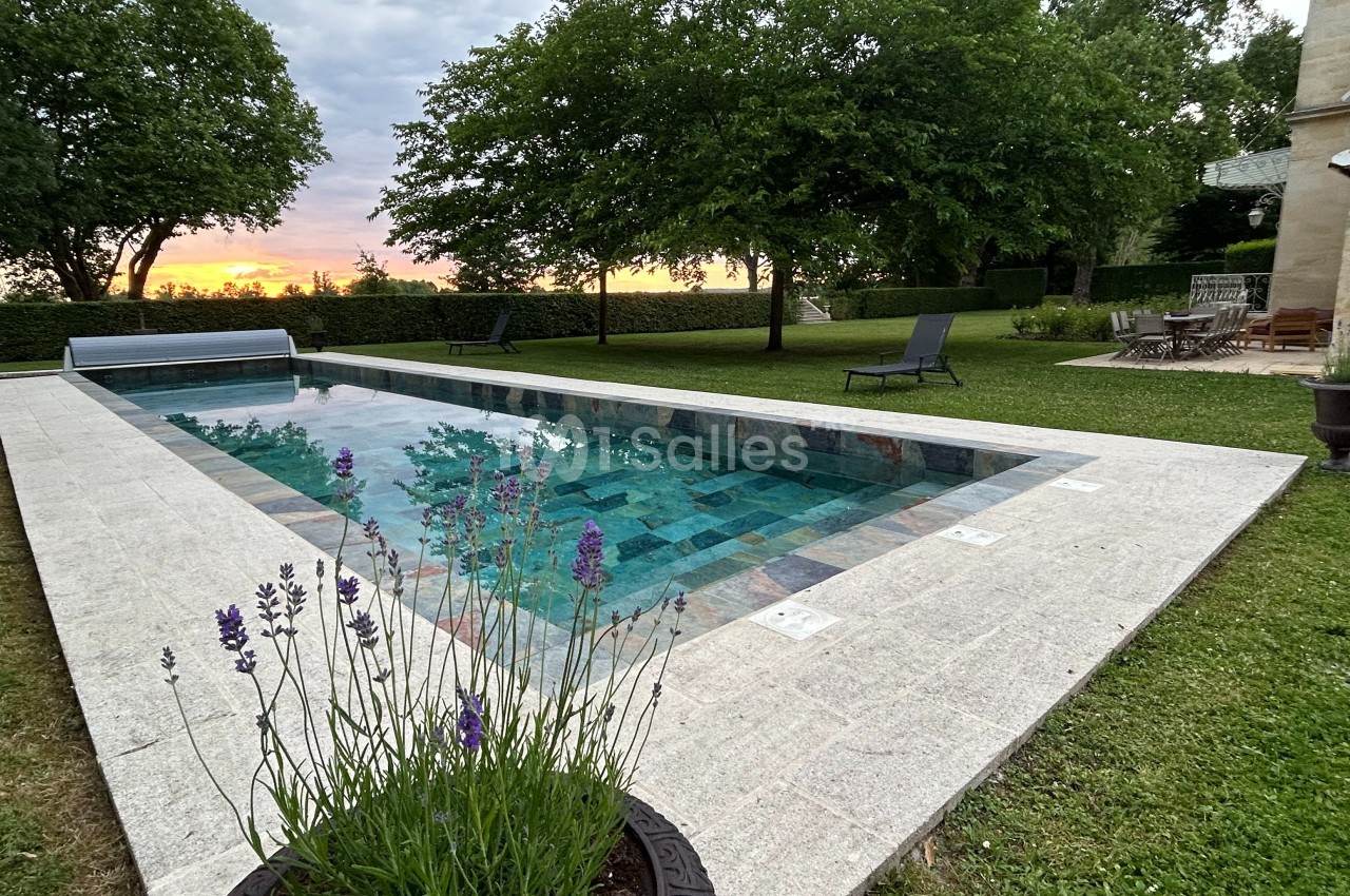 Piscine rectangulaire entourée de dalles claires, avec des arbres et un coucher de soleil en arrière-plan.