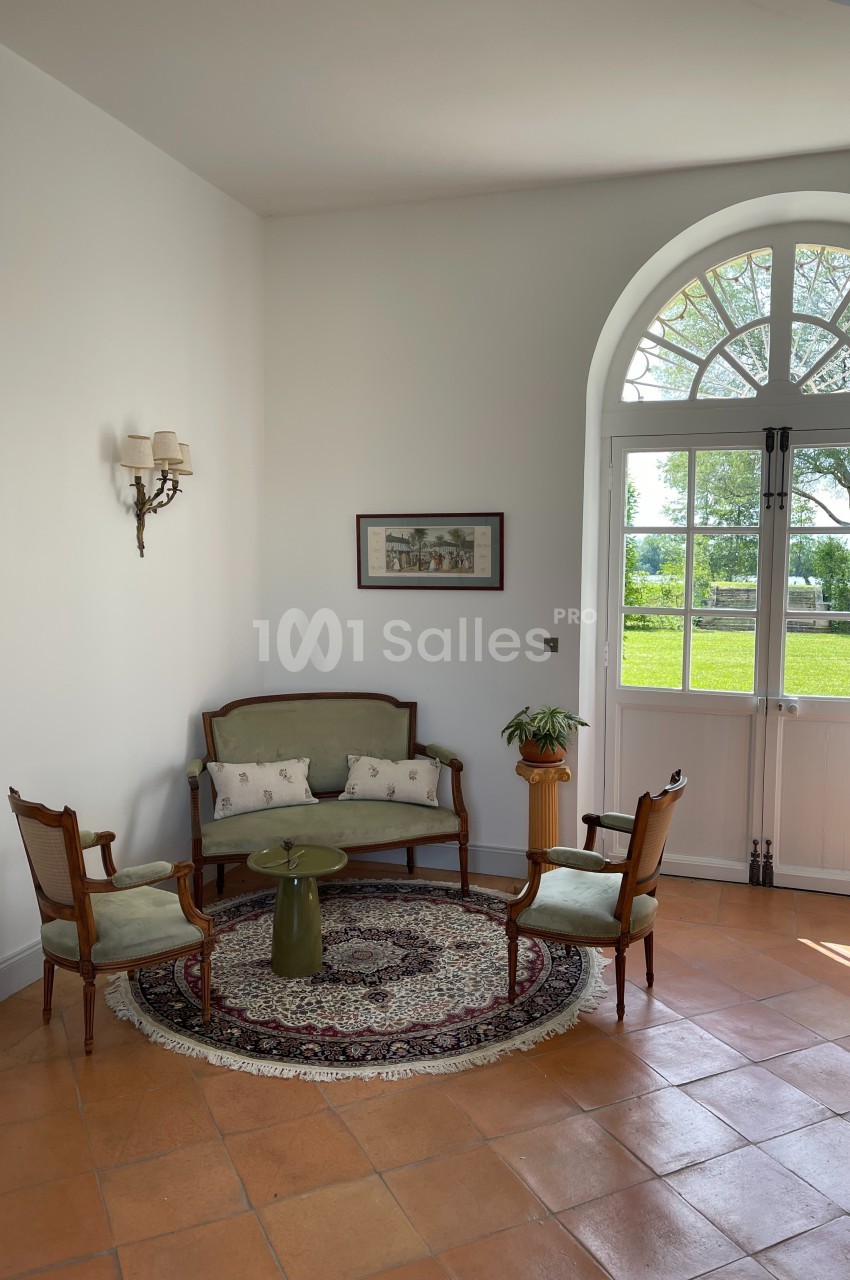 Petit salon lumineux avec canapé, deux fauteuils, table basse verte, tapis rond et vue sur un jardin par une porte vitrée.