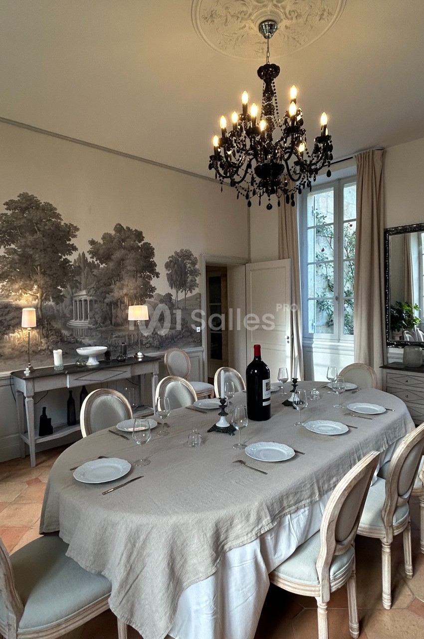 Salle à manger élégante avec table dressée, lustre noir, fresque murale et lumière naturelle entrant par une fenêtre.
