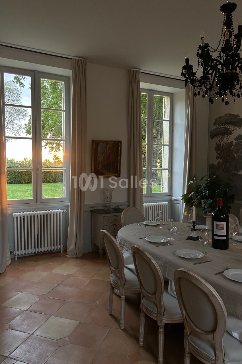 Salle à manger élégante avec table dressée, chandelier noir, grandes fenêtres et vue sur un coucher de soleil.
