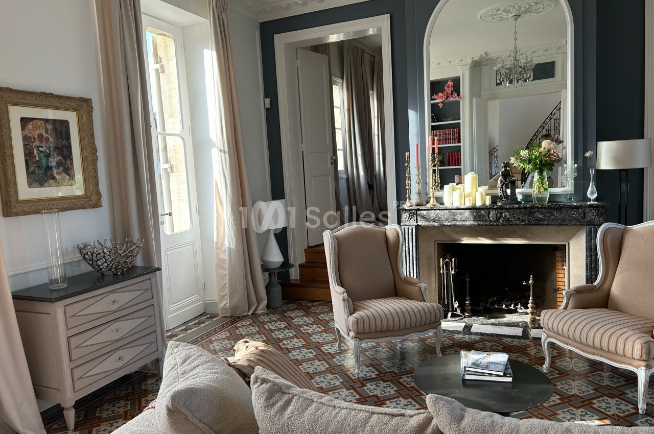 Salon élégant avec cheminée, fauteuils classiques, table basse, grandes fenêtres et carrelage décoratif au sol.