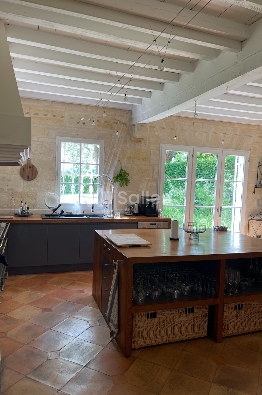 Cuisine lumineuse avec murs en pierre, grandes fenêtres, îlot central en bois et sol en carreaux de terre cuite.