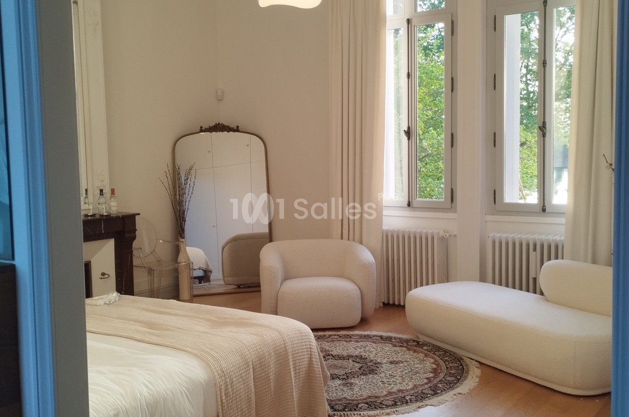 Chambre lumineuse avec lit, fauteuils blancs, miroir ancien, tapis rond et grandes fenêtres donnant sur un espace vert.