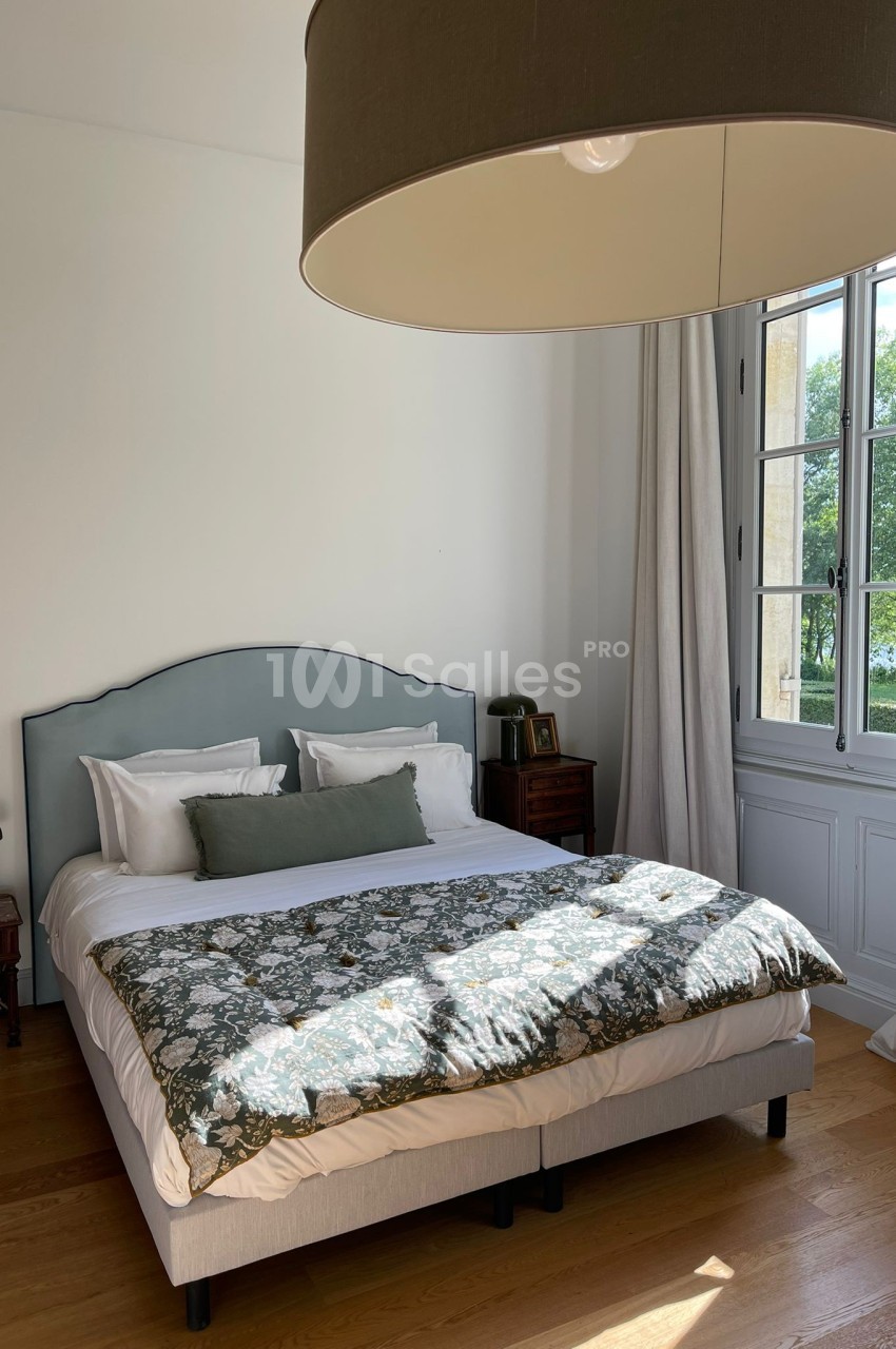 Lit double avec tête de lit bleue, couette à motifs floraux, lampe suspendue et fenêtre donnant sur un paysage verdoyant.