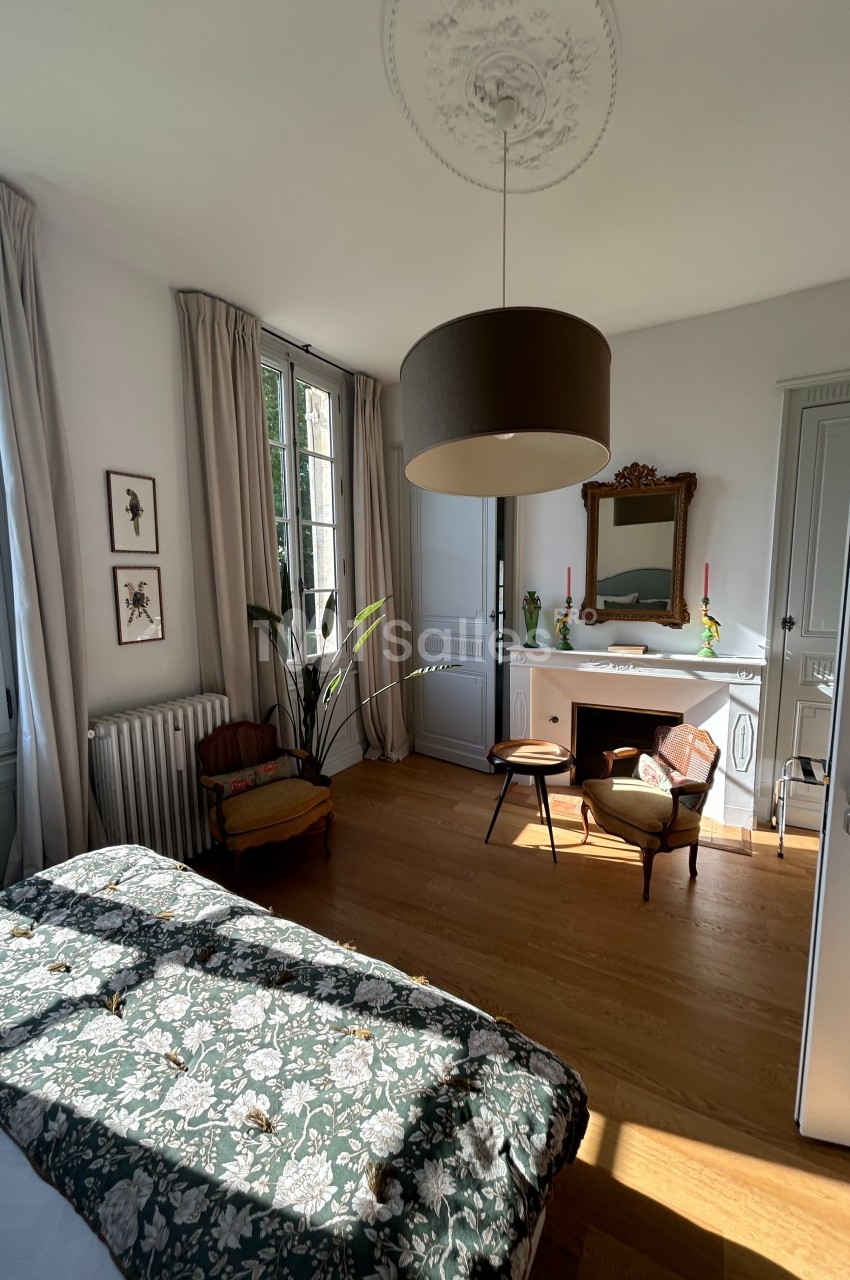 Chambre lumineuse avec lit à motif floral, parquet en bois, cheminée décorative et grandes fenêtres avec rideaux beiges.