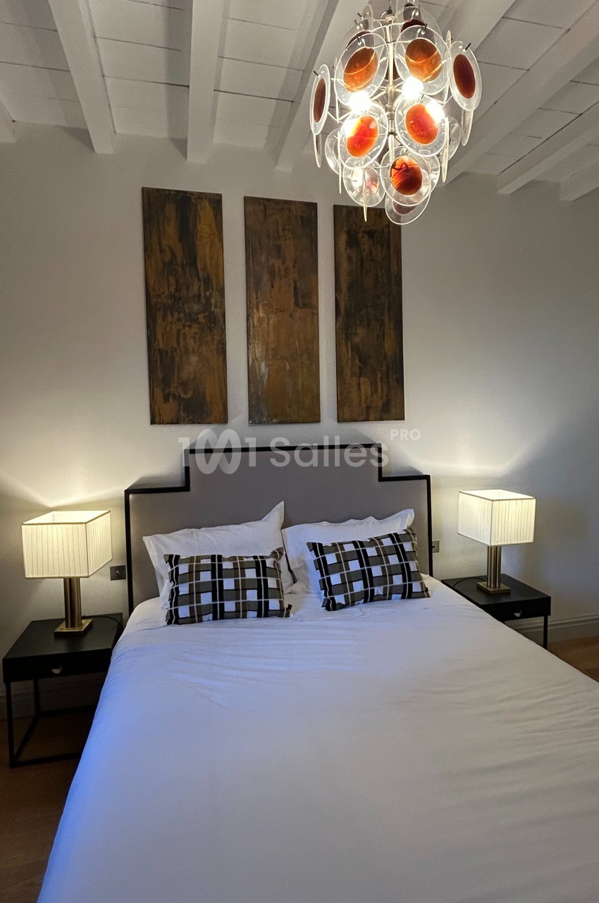 Lit double avec coussins à motifs, deux lampes de chevet allumées et un lustre moderne sous un plafond blanc.